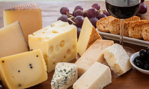 Assorted cheeses (such as ementhal, blue cheese, pecorino, parmesan and manchego among others) served with bread, grapes, black olives and a glass of red wine on a wooden table. Typical appetizer from mediterranean culture. Studio photography. High angle view. Natural day lighting. Still life. Horizontal composition.