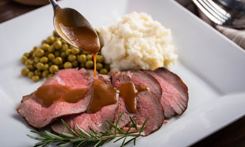 pouring gravy sauce on sliced roasted beef with mushed potato and sweet peas