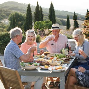 A group of mature friends are sitting around an outdoor dining table, eating and drinking. They are celebrating their holiday with a glass of champagne and enjoying each others company. The image has been taken in Tuscany, Italy.